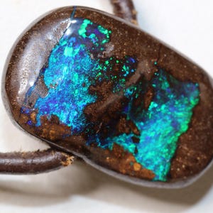 Könnte beinhalten: Ein polierter, ovaler Anhänger mit einem lebendigen Opal in einer dunkelbraunen Matrix. Der Opal zeigt ein auffälliges Farbspiel mit blauen und grünen Reflexen. Der Anhänger ist an einer braunen Lederschnur befestigt.