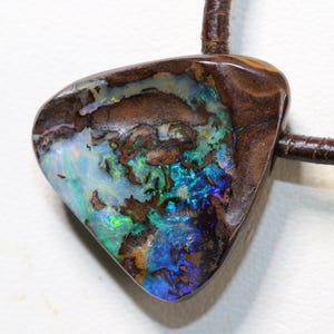 Könnte beinhalten: Ein dreieckiger Boulder-Opal-Anhänger mit einer dunkelbraunen Eisensteinmatrix und leuchtenden Blitzen aus irisierendem Blau, Grün und Weiß. Der polierte Edelstein ist an einem dunkelbraunen Lederband befestigt.