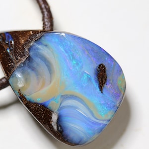 Könnte beinhalten: Ein tropfenförmiger Boulder-Opal-Anhänger mit polierter Oberfläche, der wirbelnde Muster in schillerndem Blau, Grün und Creme auf einer dunkelbraunen Eisensteinmatrix zeigt, befestigt an einer dunkelbraunen geflochtenen Lederkordel.