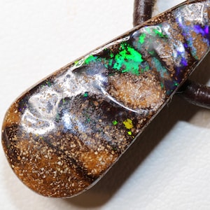 Könnte beinhalten: Tropfenförmiger Anhänger mit einem Opal. Der Opal zeigt grüne, blaue und gelbe Farbtöne auf einem dunkelbraunen Untergrund. Er ist an einer braunen Lederschnur befestigt und kann als Halskette getragen werden. Der Anhänger ist etwa 4 cm lang.