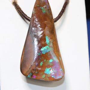 Könnte beinhalten: Ein dreieckiger Boulder-Opal-Anhänger mit einer polierten braunen Eisensteinmatrix und lebhaften, irisierenden grünen und violetten Farbspielen, der an einer dunkelbraunen Kordelkette hängt, die durch die Oberseite des Steins geführt ist.