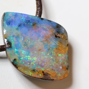 Könnte beinhalten: Ein Anhänger mit einem Opal-Edelstein, der eine Vielzahl von Farben aufweist, darunter Blau, Grün und Orange. Der Opal ist an einer dunkelbraunen Lederschnur befestigt. Der Edelstein hat eine unregelmäßige Form. Ein einzigartiges Schmuckstück.