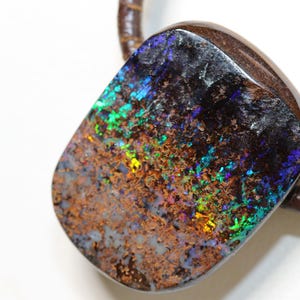 Op de afbeelding: Een gepolijste boulder-opaal hanger met een donkerbruine ijzersteenmatrix met levendige flitsen van groene, blauwe en oranje iriserende kleuren. De steen is bevestigd aan een dikke, donkerbruine gevlochten leren koordketting.