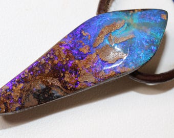Puede incluir: Un colgante de ópalo alargado con una rica exhibición de colores. La piedra presenta una base de púrpura intenso y azul, acentuada con parches marrones y toques de verde azulado. El colgante está sujeto a un cordón marrón.