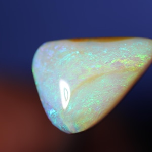 Può includere: Una gemma di opale lucidata con un colore di base bianco crema perlescente. La pietra mostra lampi iridescenti di verde, blu e giallo. L'opale ha una forma liscia e arrotondata ed è ambientata su uno sfondo blu scuro e marrone.