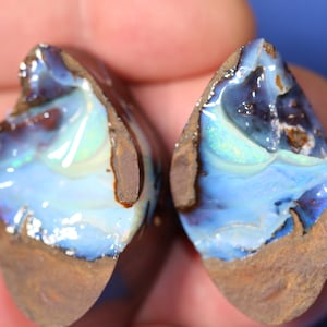 Puede incluir: Dos piedras de ópalo natural en bruto con un brillo iridiscente azul, verde y blanco. Las piedras están engastadas en una matriz marrón oscuro.