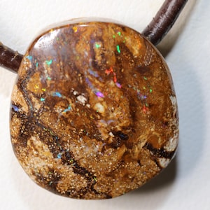 May include: An oval pendant with a brown and tan colour scheme. The stone displays iridescent flashes of colour, including red, green, and blue. It is attached to a dark brown leather cord, creating a natural and earthy aesthetic.