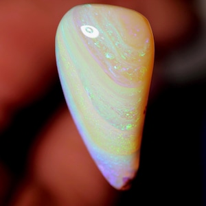 Op de afbeelding: Een gepolijste opaal edelsteen met een druppelvorm. De steen vertoont een wervelend patroon van kleuren, waaronder wit, geel, groen en hints van paars. De opaal wordt tegen een vage achtergrond gehouden.