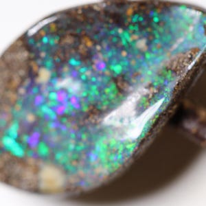 Könnte beinhalten: Ein dunkelbrauner und schwarzer Opal mit einem grün-blau-violetten, schillernden Glanz. Der Opal ist in einer rauen, natürlichen Fassung gefasst.