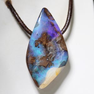 Könnte beinhalten: Ein Anhänger mit einem polierten, unregelmäßig geformten Boulder-Opal mit leuchtenden Blau-, Violett- und Grüntönen, befestigt an einem dunkelbraunen Lederband. Der Stein zeigt natürliche braune Matrixeinschlüsse und eine glatte, glänzende Oberfläche.
