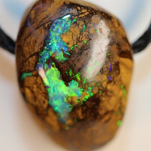 Könnte beinhalten: Ein dunkelbrauner und schwarzer Opal-Anhänger mit grün-blauen, schillernden Blitzen. Der Opal ist in silberfarbenem Metall gefasst und hängt an einer schwarzen Schnur.