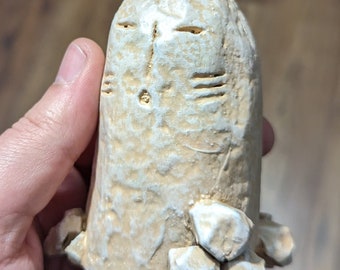 May include: A beige, hand-carved sculpture with a stylized face and a textured surface. The sculpture has a cylindrical shape with a base adorned with small, white stone-like elements. The face features carved eyes, a mouth, and a small decorative element.
