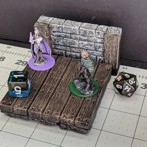 May include: A miniature tabletop gaming set with a stone wall backdrop, a wooden platform, and two miniature figures. The set includes a purple base with a miniature figure of a person in a purple robe holding a staff, a green base with a miniature figure of a person in a green cloak, and two dice. One die is blue and shows the number 1, and the other die is brown and shows the numbers 14, 8, and 20.