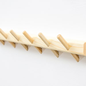 Puede incluir: Un perchero de pared de madera con siete ganchos angulados. El perchero está hecho de madera de color claro y tiene un acabado natural.