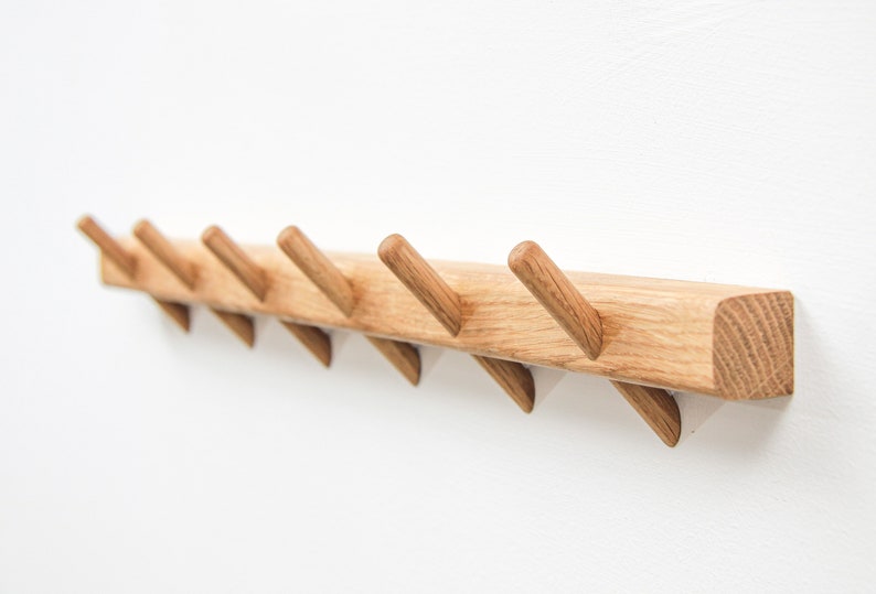 Op de afbeelding: Een houten wandbevestigde kapstok met zes schuin geplaatste haken. De kapstok is gemaakt van natuurlijk hout met een lichtbruine afwerking.