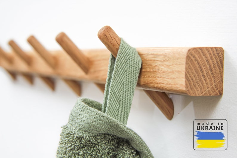 Op de afbeelding: Een houten wandbevestigde kapstok met zes haken. De kapstok is gemaakt van lichtbruin hout en heeft een natuurlijke afwerking. Een groene handdoek hangt aan een van de haken. De kapstok heeft de tekst "made in UKRAINE" op de muur eronder.