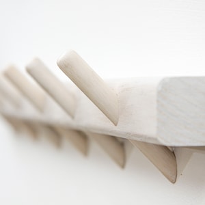Puede incluir: Perchero de pared de madera blanco con múltiples ganchos. El perchero está hecho de madera sin terminar y tiene un diseño simple y moderno.