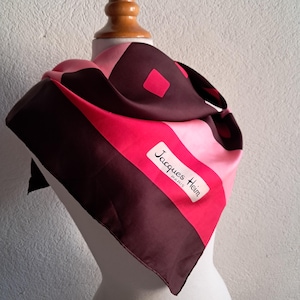 May include: A silk scarf with a geometric design in shades of pink, red, and brown. The scarf features the text "Jacques Heim PARIS" on a white label. The scarf is draped over a mannequin.