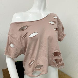 Op de afbeelding: Een stofroze off-the-shoulder T-shirt met meerdere ovale uitsparingen. De top met korte mouwen wordt op een mannequin gepresenteerd. De uitsparingen zijn gelijkmatig verdeeld over de voorkant en de mouwen van het shirt.