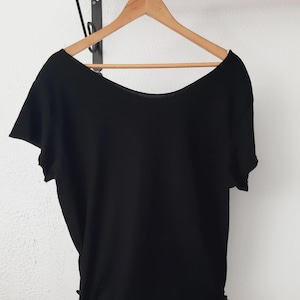 Op de afbeelding: Een zwarte top met korte mouwen en een brede, ronde hals hangt aan een houten kleerhanger. De top heeft een geplooid detail aan de zijkant en wordt gepresenteerd tegen een witte muur. De top is effen van kleur en lijkt van een zacht materiaal te zijn.