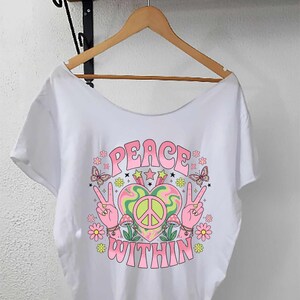 Könnte beinhalten: Ein weißes T-Shirt mit einem Grafikdruck, der den Text "PEACE WITHIN" in rosa Buchstaben zeigt. Das Design umfasst zwei Hände, die Friedenszeichen formen, ein zentrales Herz mit einem Friedenssymbol, Pilze, Schmetterlinge und kleine Blumen.