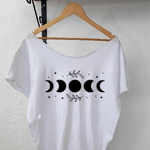 Könnte beinhalten: Weißes T-Shirt mit Rundhalsausschnitt und kurzen Ärmeln. Auf der Vorderseite befindet sich eine schwarze Grafik der Mondphasen mit Sternen und Blättern. Das T-Shirt hängt an einem Holzbügel.