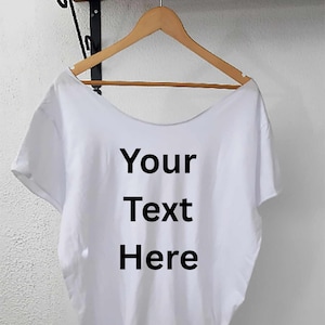 以下が含まれることがあります： 「Your Text Here」の黒い文字が入った白いTシャツが、木製のハンガーに掛けられています。Tシャツは丸首で半袖です。背景は無地の白い壁です。