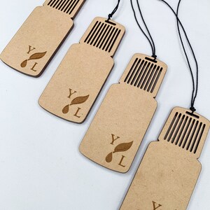 May include: Four wooden essential oil diffuser tags with black string. Each tag is shaped like a bottle with a slotted top and features the letters "YL" with a leaf and drop design. The tags are light brown and are arranged diagonally.