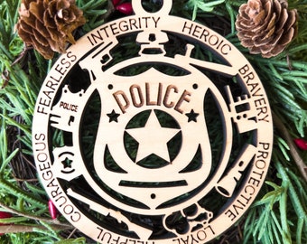 Könnte beinhalten: Ein hölzerner Polizei-Abzeichen-Weihnachtsschmuck mit schwarzem Band. Der Schmuck zeigt das Wort "POLIZEI" und andere Wörter wie "INTEGRITÄT" und "HEROISCH". Umgeben von Tannennadeln, Tannenzapfen und roten Beeren.