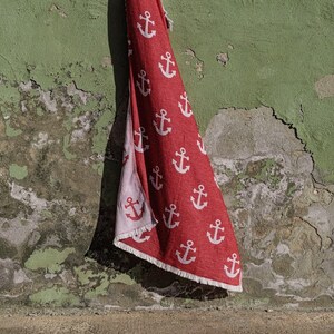 Könnte beinhalten: Ein rotes Strandtuch mit weißen Ankermotiven hängt an einer grünen Wand. Die Wand ist strukturiert und weist einige abblätternde Farbe auf.