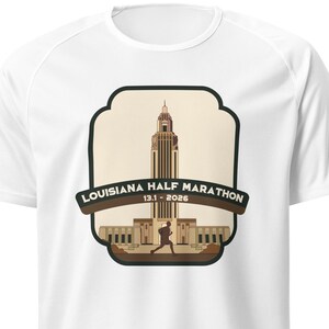 May include: White running shirt with a brown graphic of a building and a runner. The graphic says "Louisiana Half Marathon 13.1 - 2026".