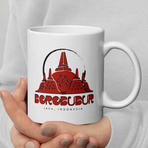 Puede incluir: Taza de cerámica blanca con una ilustración roja y negra del templo de Borobudur en Java, Indonesia. El texto "BOROBUDUR JAVA, INDONESIA" está impreso debajo de la imagen.