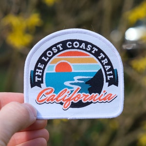 Peut inclure: Écusson blanc avec un graphique coloré d'un coucher de soleil sur une côte. Le texte "THE LOST COAST TRAIL California" est imprimé sur l'écusson.