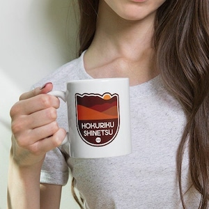 Puede incluir: Taza de cerámica blanca con un diseño gráfico con el texto "HOKURIKU SHINETSU" en un escudo. El diseño incluye una cordillera, un sol poniente y un pequeño círculo. La taza tiene un asa curva y la sostiene una persona.