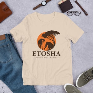 Peut inclure: Un t-shirt beige avec une silhouette brune d'une girafe et d'un éléphant debout sous un arbre devant un soleil orange. Le texte "ETOSHA National Park - Namibia" est imprimé sous l'image.