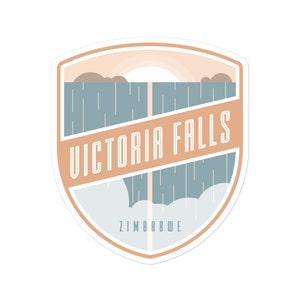 May include: A tan and blue shield-shaped sticker with the text "VICTORIA FALLS" in white outlined letters. The sticker also includes a stylized image of Victoria Falls with a rainbow above it and the text "ZIMBABWE" at the bottom.
