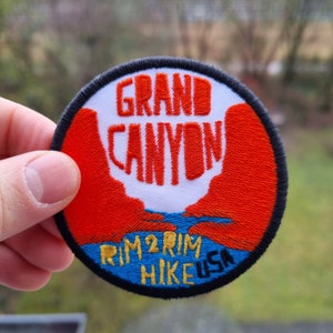 May include: Round embroidered patch with the words "GRAND CANYON" in orange, over a red and blue landscape design. The text "RIM 2 RIM HIKE USA" is below the canyon illustration. The patch has a black border.