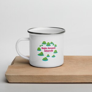 Könnte beinhalten: Eine weiße Emailletasse mit silbernem Rand. Die Tasse zeigt eine Grafik mit grünen Inseln, blauem Wasser und dem Text "Raja Ampat Islands" in rosa und weißen Buchstaben. Der Text "INDONESIA" steht in kleineren weißen Buchstaben unter dem Haupttext.