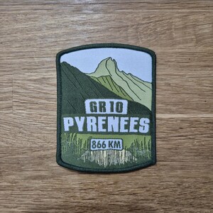 Puede incluir: Parche bordado verde y blanco con el texto "GR10 PYRENEES" y "866 KM". El parche presenta un pico de montaña y un campo verde.