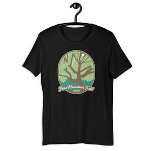Puede incluir: Camiseta negra con una ilustración verde y marrón de un árbol en un círculo con el texto "Pine Mountain Trail" debajo.