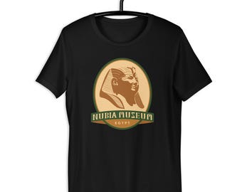 May include: Black t-shirt featuring a circular design with a pharaoh's profile in brown and tan, encircled by a green border. The text "NUBIA MUSEUM EGYPT" is displayed below the image in green and tan.
