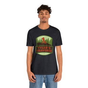 May include: Black t-shirt featuring a graphic with the text "TYLER'S TRAVERSE" in red and white, with a mountain biker silhouette. The design includes a green and brown landscape with trees, and the text "SUNRIVER | OR".
