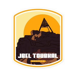 Puede incluir: Una pegatina ilustrada amarilla y naranja con un pico de montaña y un pequeño edificio en la cima. El texto "Jbel Toubkal" y "MOROCCO" están impresos en la pegatina.