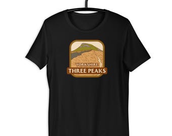 May include: Black t-shirt with a graphic of a mountain scene and the text "YORKSHIRE THREE PEAKS" in a brown frame. The design incorporates green, tan, and brown hues. The t-shirt is displayed on a black hanger.