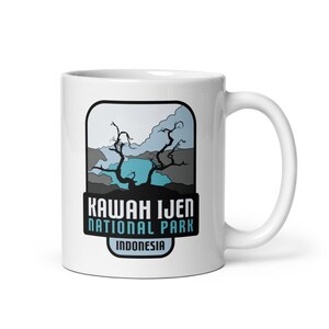 Może przedstawiać: Biały ceramiczny kubek z grafiką Parku Narodowego Kawah Ijen w Indonezji. Projekt zawiera krajobraz z turkusowym jeziorem, nagimi drzewami oraz napisem "KAWAH IJEN NATIONAL PARK INDONESIA" w kolorze czarnym i jasnoniebieskim.