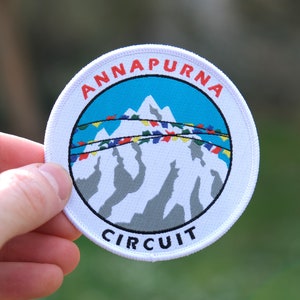 May include: White round patch with the text "Annapurna Circuit" in red and blue lettering. The patch features a blue sky with a mountain range and colorful prayer flags.