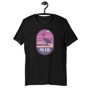 Może przedstawiać: Czarny t-shirt z grafiką sylwetki ptaka w krajobrazie oraz napisem "PALO VERDE NATIONAL PARK, COSTA RICA" w fioletowo-różowym owalnym wzorze. Koszulka wisi na czarnym wieszaku.