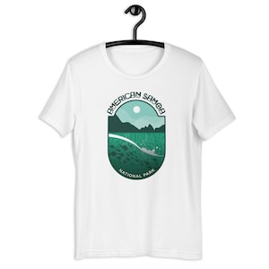 Könnte beinhalten: Weißes T-Shirt mit einer Grafik des American Samoa National Park. Das Design zeigt eine türkis-grüne Landschaft mit dem Text "AMERICAN SAMOA NATIONAL PARK" in kreisförmiger Anordnung. Das T-Shirt hängt an einem schwarzen Kleiderbügel.