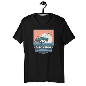 Może przedstawiać: Czarny t-shirt z grafiką drzewa, słońca i ptaków oraz napisem "PALO VERDE NATIONAL PARK Costa Rica". Projekt jest w odcieniach niebieskiego, zielonego, pomarańczowego i białego. Koszulka wisi na czarnym wieszaku.