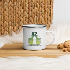 Könnte beinhalten: Weißer Emaillebecher mit silbernem Rand und Henkel. Der Becher zeigt eine grün-weiße Grafik eines Leuchtturms mit dem Text "E7 EUROPEAN LONG DISTANCE PATH". Der Becher steht auf einer Holzoberfläche mit Keksen.